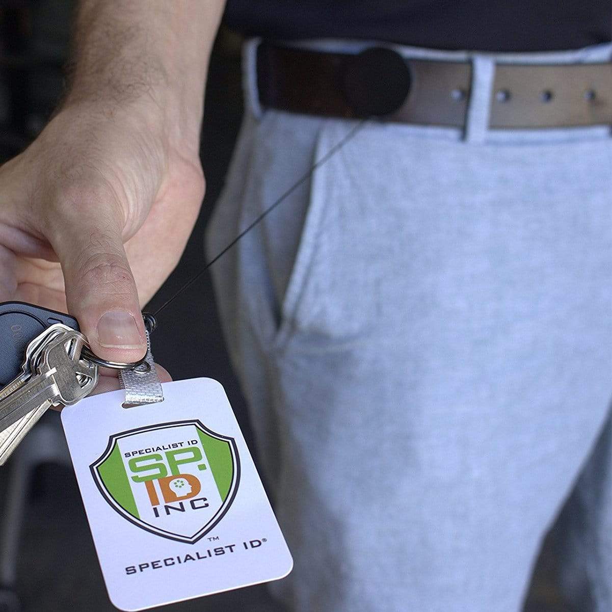 A person holding a Black Chrome Heavy Duty Badge Reel with Belt Clip (2120-3300) with keys attached, featuring a badge with a "Specialist ID Inc" logo. The retractable reel is clipped to the belt clip on their brown belt. They are wearing light gray pants.