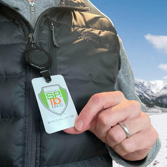 Person wearing a black puffer vest and gray hoodie, displaying a badge from a retractable lanyard with the "Specialist ID" logo, standing outdoors in a snowy mountain landscape. The Ski Pass and Sports Retractable Reel (P/N 2120-4001)'s rugged plastic design ensures durability in harsh conditions.