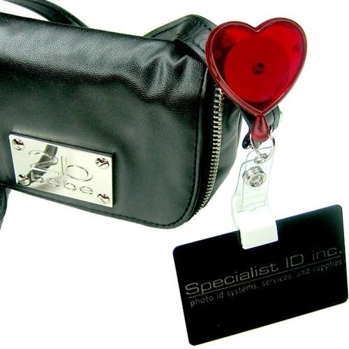 A black ID holder with "Specialist ID Inc." text is attached to a black bag by a Heart Shaped Badge Reel With Rotating Spring Clip (P/N 2120-761X) with a retractable cord.