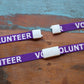 Two Purple "Volunteer" Breakaway Lanyards W/ Swivel Hook 2138-5230 with the word "VOLUNTEER" in white text, placed on a wooden surface with a blue paint patch in the background. Each lanyard features a white plastic clasp.
