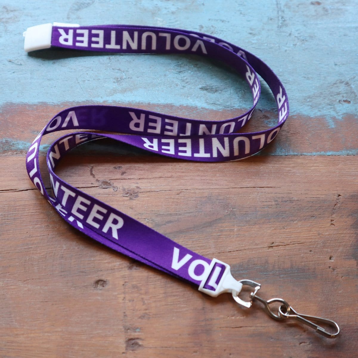 A purple "Volunteer" Breakaway Lanyard W/ Swivel Hook 2138-5230 with white text reading "VOLUNTEER" repeatedly, featuring a swivel hook at the bottom. The lanyard is laid out on a worn wooden surface.