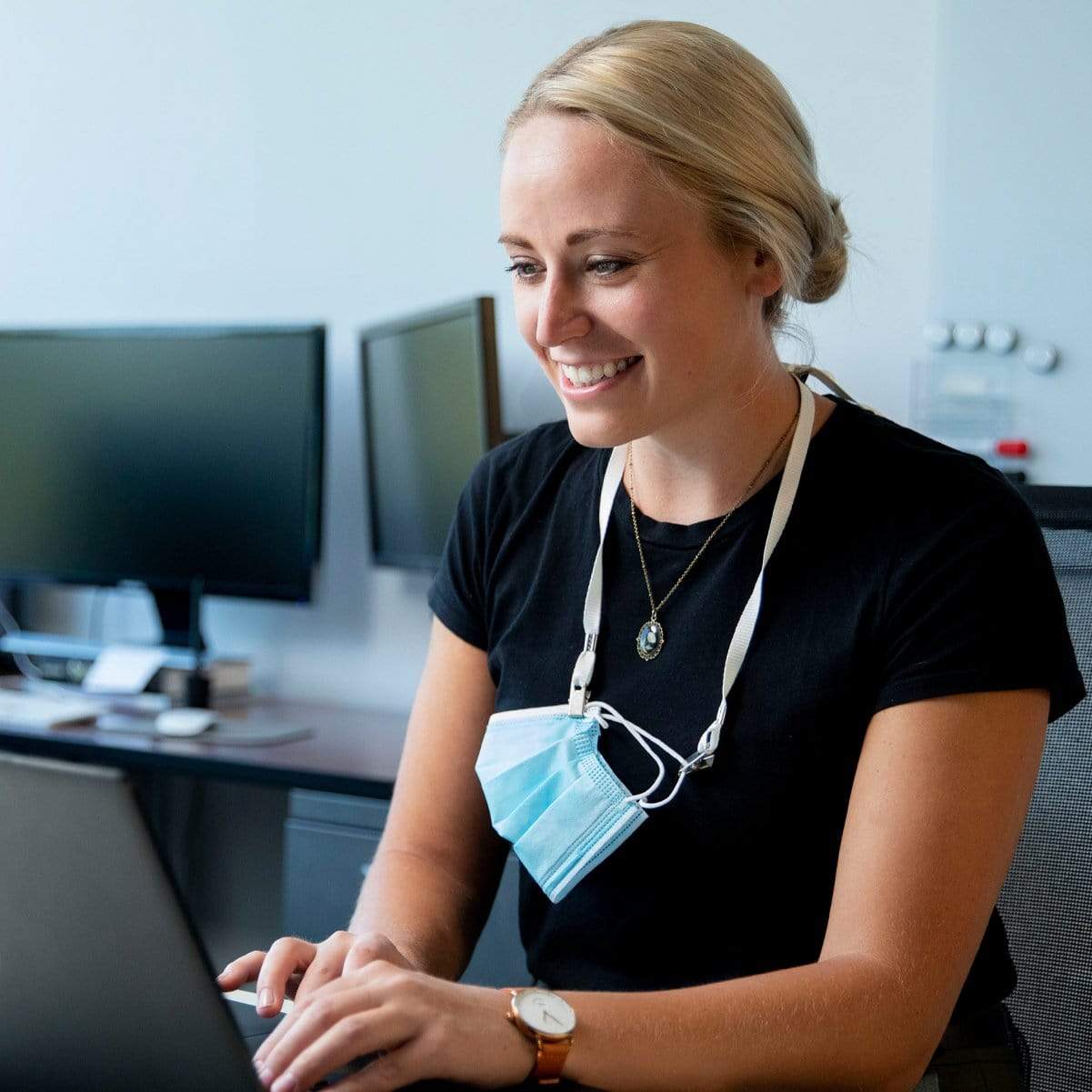 A woman wearing a black shirt and a Double Clip Lanyard with 2 Bulldog Clips - Flat, Soft Material Neck Straps for Large Badge Holder Credentials (2140-530X) with a face mask is smiling while typing on a laptop in an office setting with multiple monitors in the background.