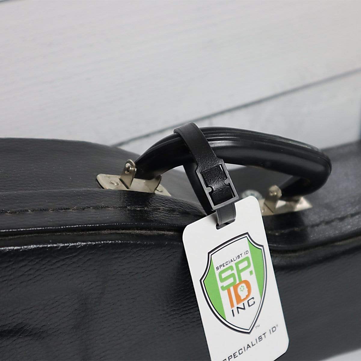 A black leather briefcase with a Plastic Luggage Strap with Post and Notch -Textured (2430-201X) attached to the handle and a durable travel strap for easy carrying. The tag features a logo with a shield and text "SIID INC Specialist ID." The briefcase is placed against a white wall.