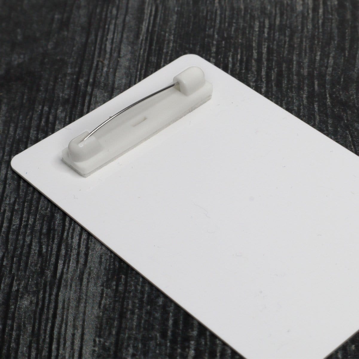 A plain white ID badge with a Safety Locking Adhesive Bar Pin - Badge & Craft Accessory 6920-3655 is placed on a dark, textured surface, showcasing its practical design as a badge and craft accessory.