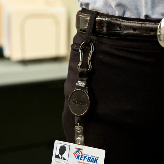A person wearing black-belted pants has a KEY-BAK Mid Size Retractable ID Carabiner (6CID) clipped to the belt loop, displaying an ID badge with a key ring attached.