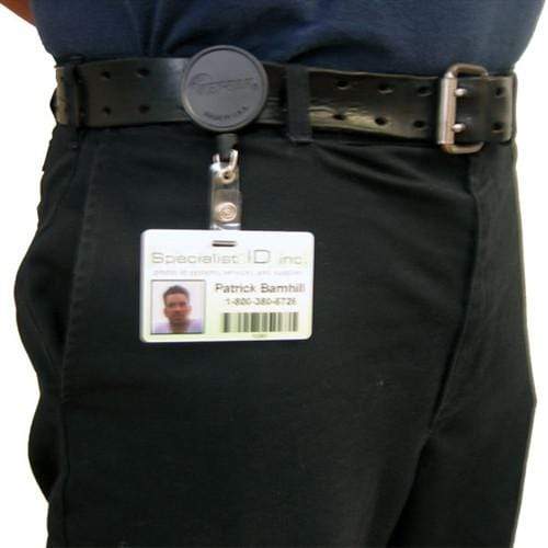 A person wearing black pants and a blue shirt has a work ID badge attached to their belt with a Key-Bak Mid Size ID Badge Reel with Steel Belt Clip (6ID). The ID includes a photo, barcode, and the name Patrick Banbali.