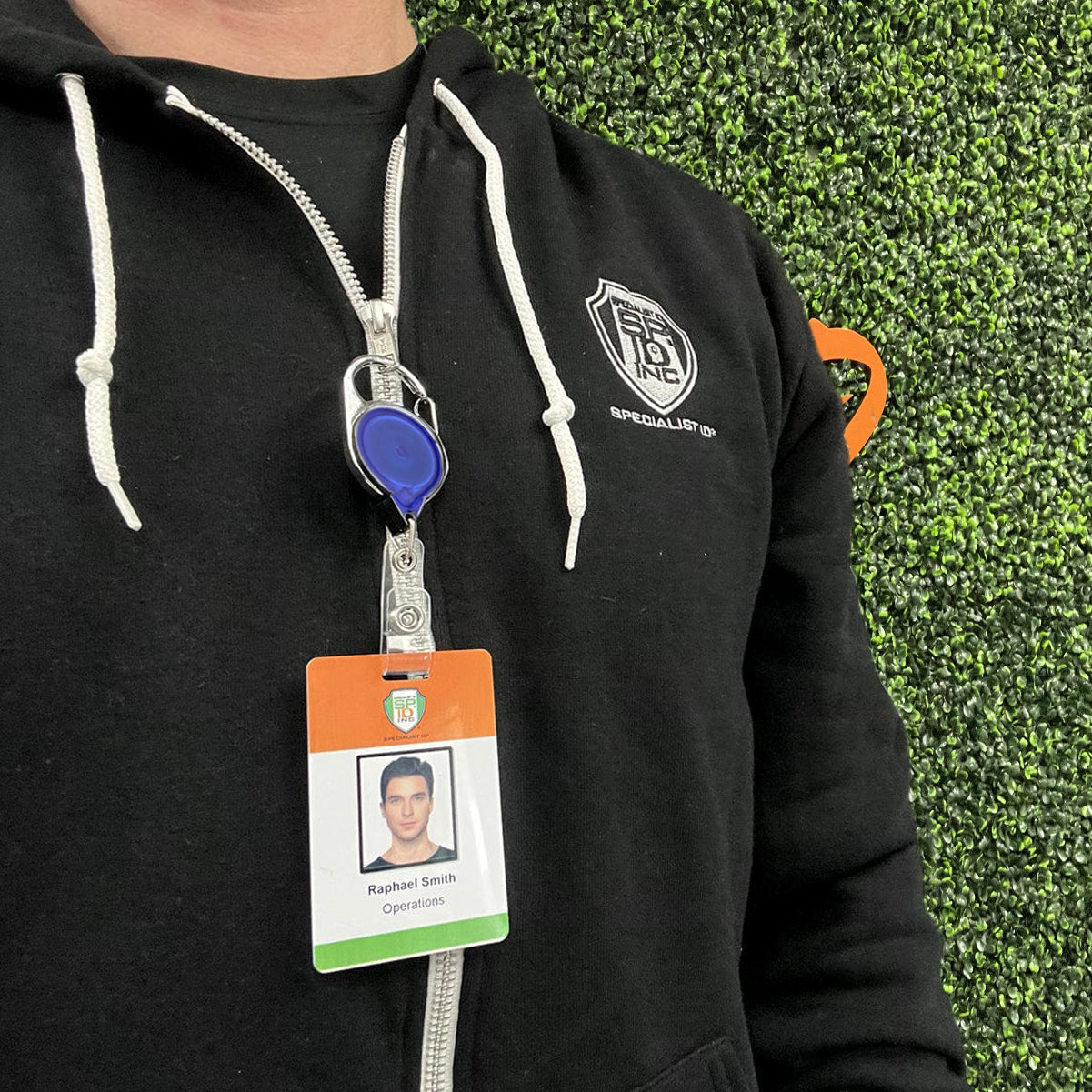 A person wearing a black zip-up hoodie with a badge that shows a photo, name, and the label "Operations," standing against a background of green foliage, has their ID secured with a Translucent Color Carabiner No Twist Badge Reel With Standard Strap Clip 704-TR.