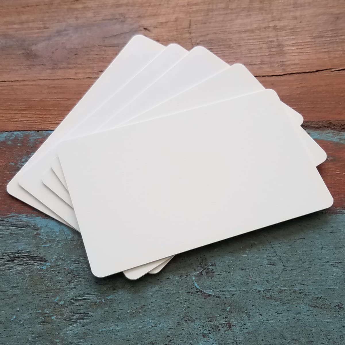A stack of Premium Blank PVC Cards for ID Badge Printers - Graphic Quality CR80 30 Mil (CR80), blank and white, fanned out on a wooden surface with a weathered texture.