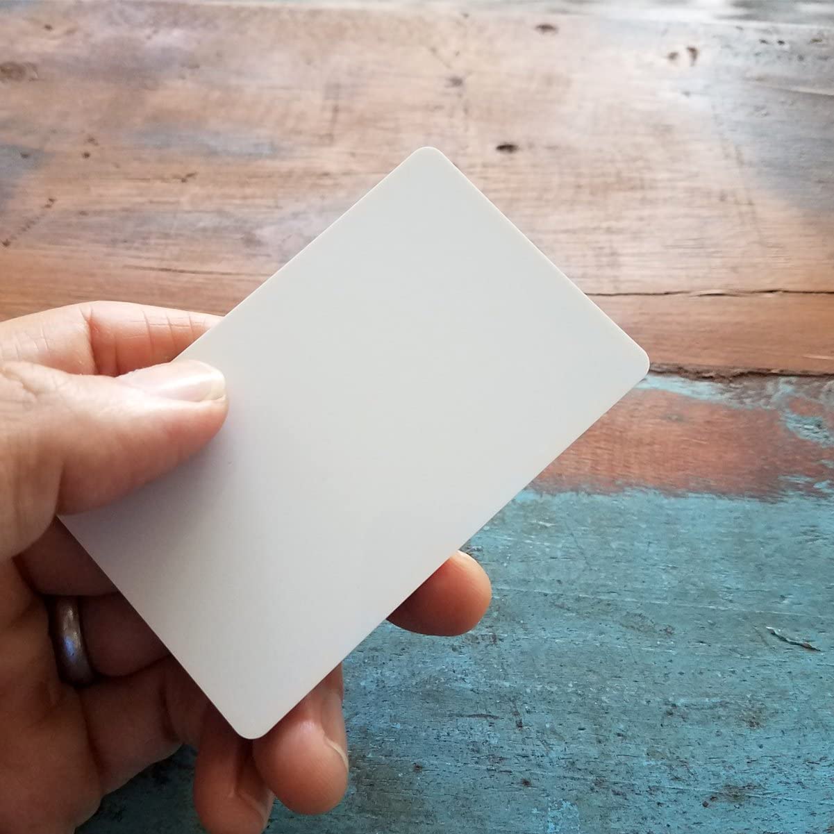 A hand holding a blank white card, perfect for Premium Blank PVC Cards for ID Badge Printers - Graphic Quality CR80 30 Mil (CR80) used in employee ID badges, against a background of wooden table with worn blue and brown paint.