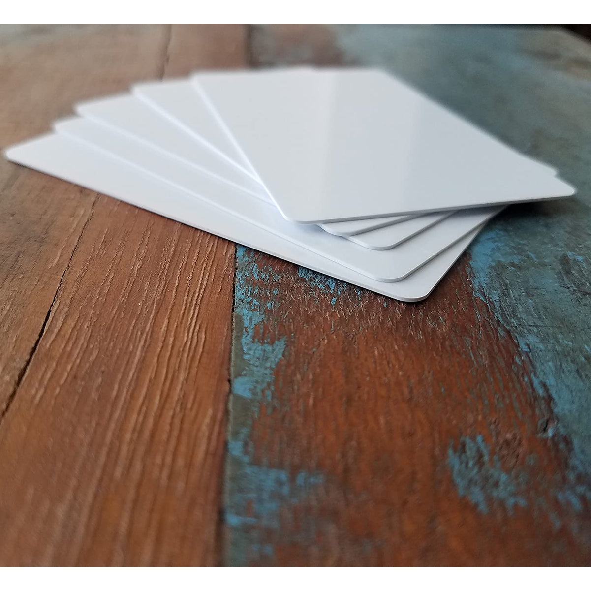 A stack of five Premium Blank PVC Cards for ID Badge Printers - Graphic Quality CR80 30 Mil (CR80) is placed on a rustic wooden surface with a mix of brown and blue paint.