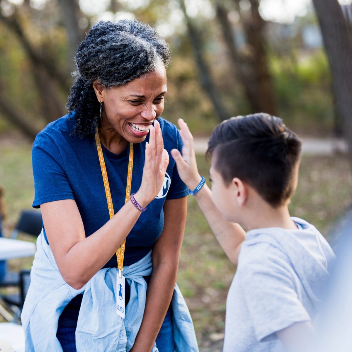 A woman and a boy share a high-five outdoors. The woman, smiling and wearing a blue shirt with an Orange Printed Bully Prevention Lanyard With Plastic Hook (P/N 2138-5240), appears encouraging. Trees are visible in the background.