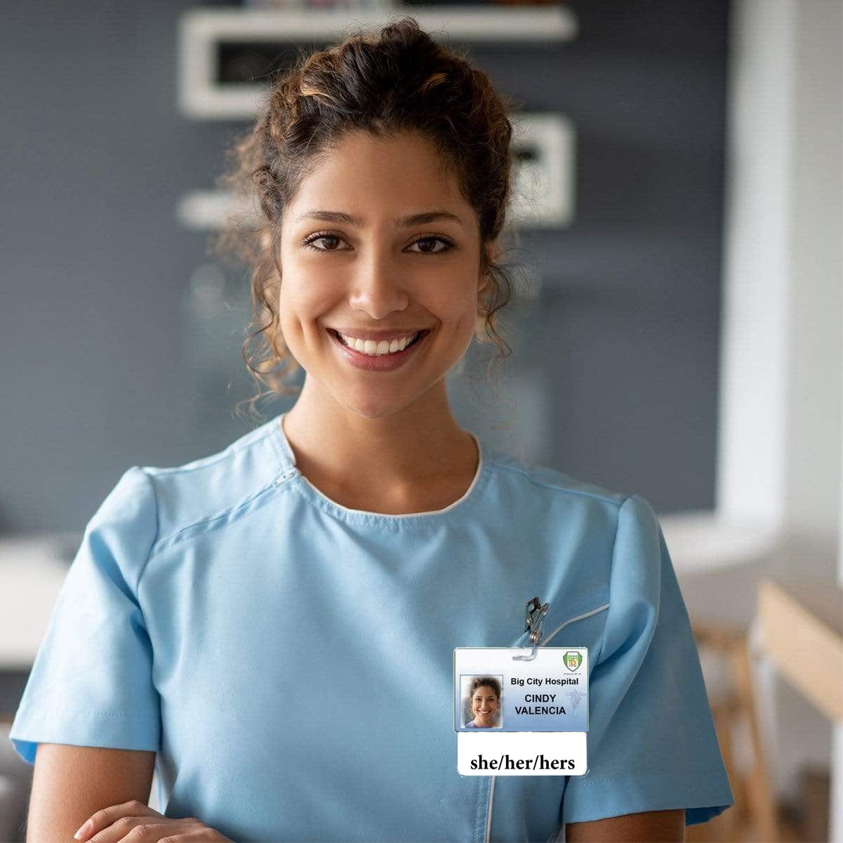 A woman with curly hair smiles while wearing light blue scrubs and an ID badge that reads "Big City Hospital, Cindy Valencia, she/her/hers," proudly displaying her Pronoun Badge Buddy She/Her/Hers Horizontal with White Border.