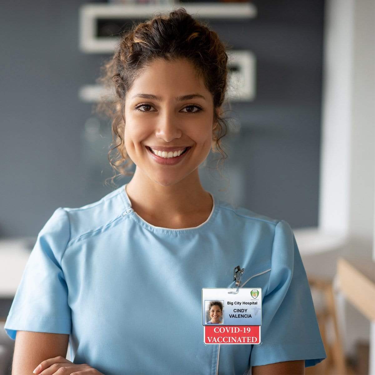 A smiling nurse in blue scrubs, wearing an ID badge that reads "Cindy Valencia," with a COVID-19 VACCINATED Badge Buddy - Horizontal Badge Backer with Red Border. The nurse's cheerful demeanor showcases a positive work environment.
