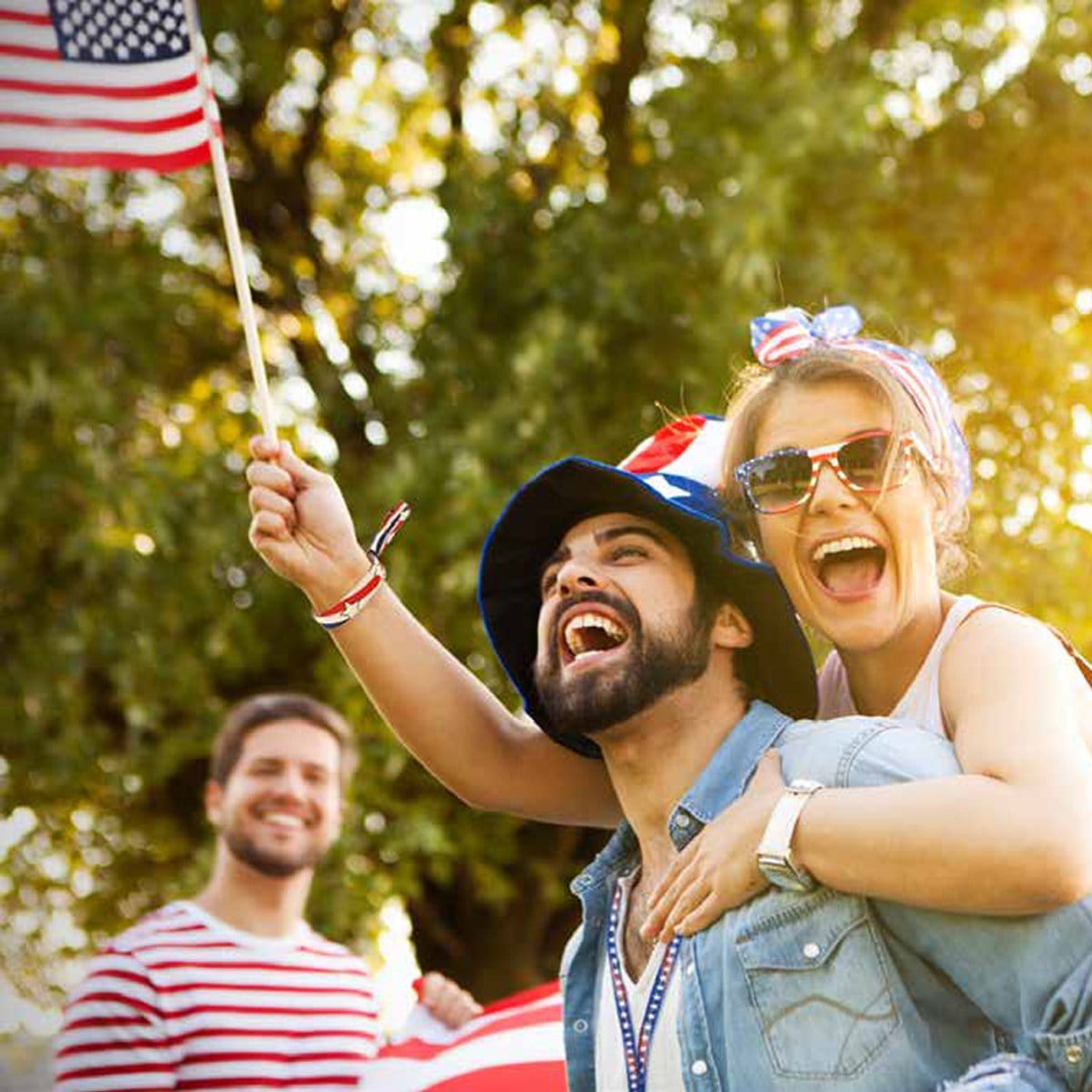 Two cheerful people celebrate outdoors, one giving a piggyback ride while holding a small American flag. A third person in a striped shirt smiles in the background, sporting colorful **100 Pack - Liberty Design 1/2" High Security Adjustable Closure Woven Wristbands**. Trees provide a lush backdrop to their joyful gathering.
