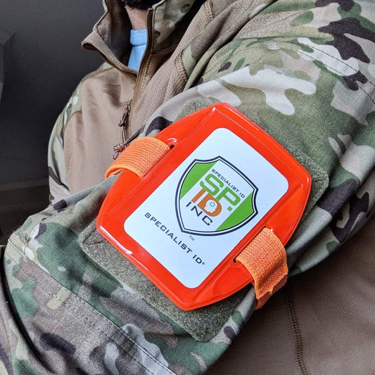 A person wearing a camouflage jacket has an orange, reflective armband with an adjustable strap and a label displaying the logo of "Vertical Reflective Armband ID Holder (R504-ARN).