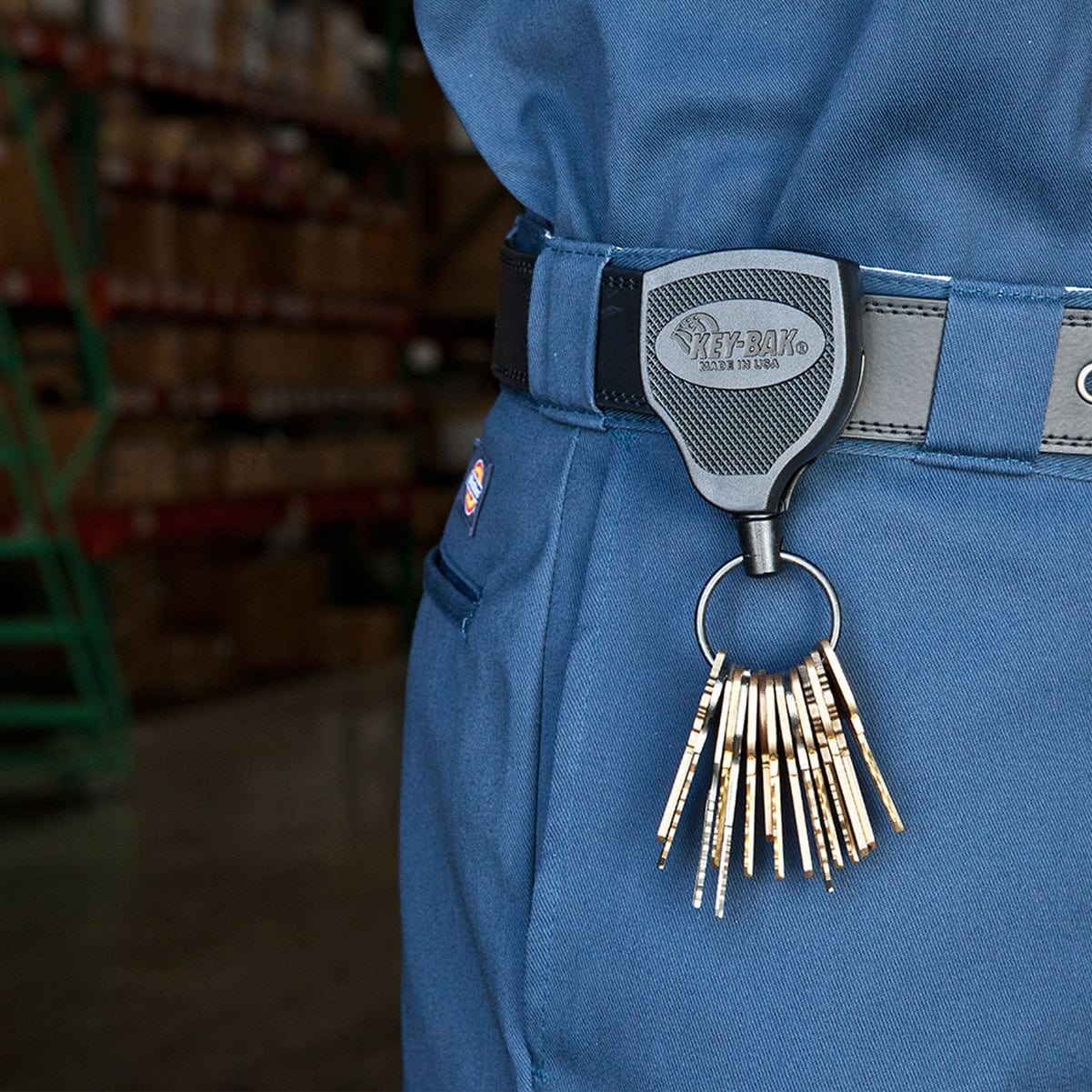 A close-up image of a person wearing a blue jumpsuit, showing a Key-Bak Super 48 Heavy Duty Key Reel with Belt Clip (S48K) with a kevlar cord attached to their belt clip, holding multiple keys. The background features stacked goods in a warehouse setting.