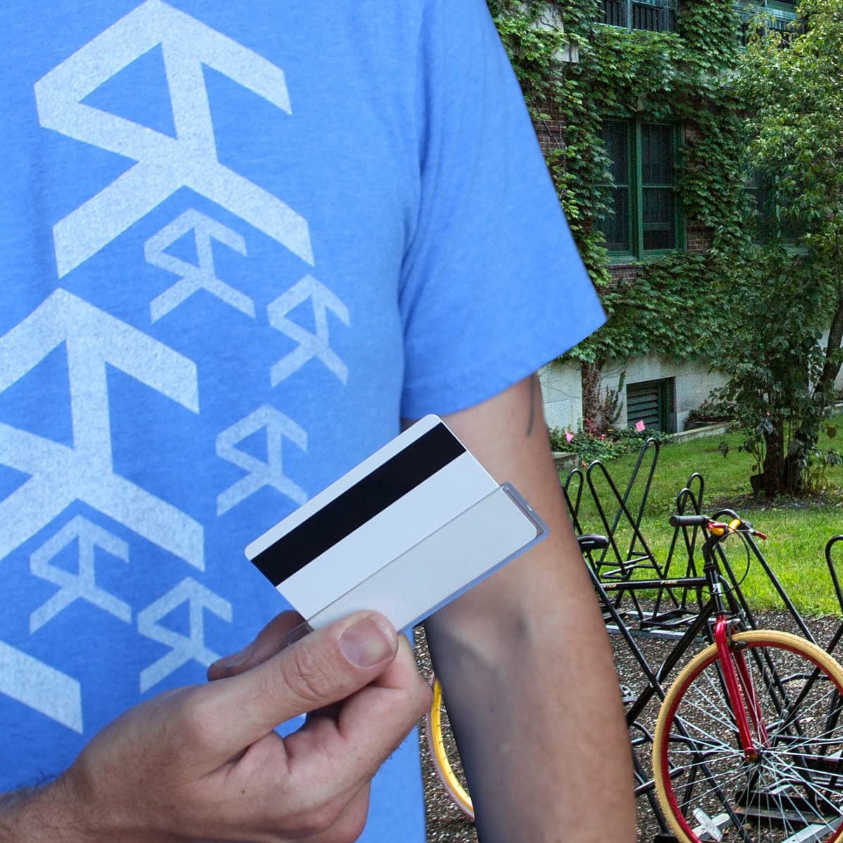 A person in a blue shirt is holding a Vertical Half Card Holder for Magnetic Stripe Swipe Cards - Heavy Duty Gripper (SPID-1380). In the background, there is a bike rack and a building covered with green vines.