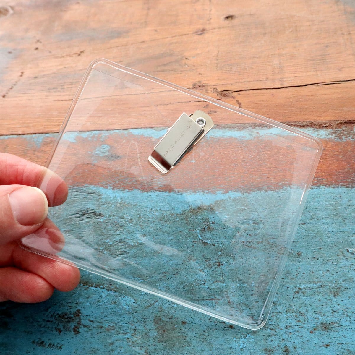 A hand holding a 4 x 3 Horizontal Event Badge Holder with Clothing Friendly Bulldog Clip - Clear Vinyl, Horizontal & Durable (SPID-1440) against a wooden surface with blue paint.