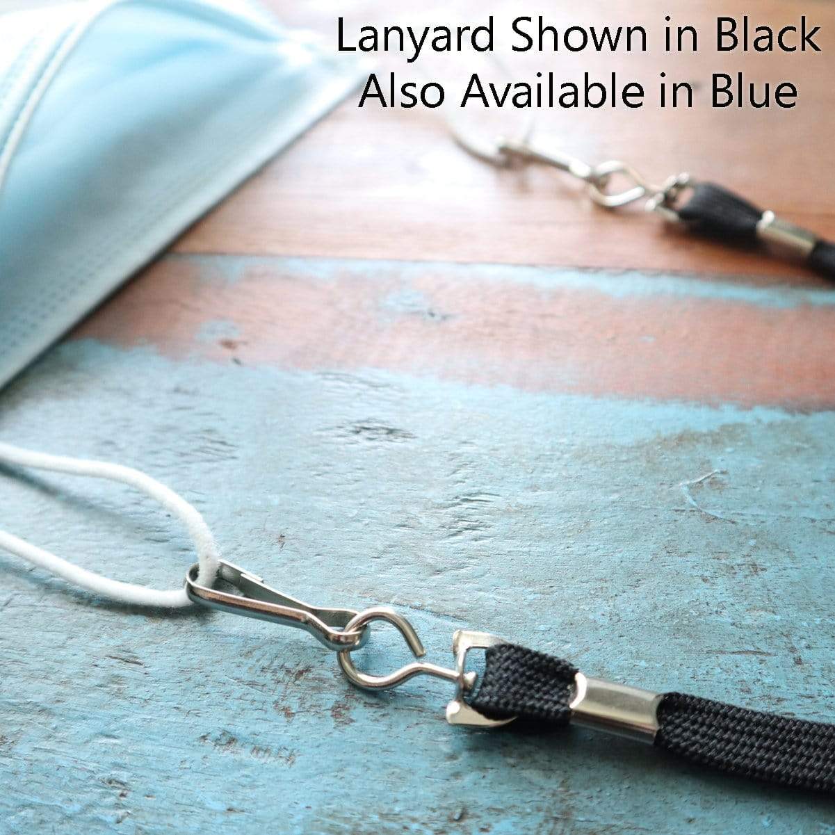 Close-up image of two lanyards, one black and one blue, attached to face masks on a wooden surface. Text reads: "Face Mask Lanyard / Hanger with Safety Breakaway Clasp - Ear Saver with J Hooks Adult Size Shown in Black Also Available in Blue." Features safety breakaway clasp for added security.