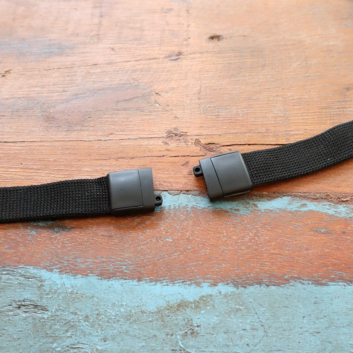 A broken black strap with a plastic buckle, resembling a Lanyard with Pen and ID Badge Holder - All in One Neck Lanyards (SPID-2380), is laid out on a wooden surface with chipped teal paint.