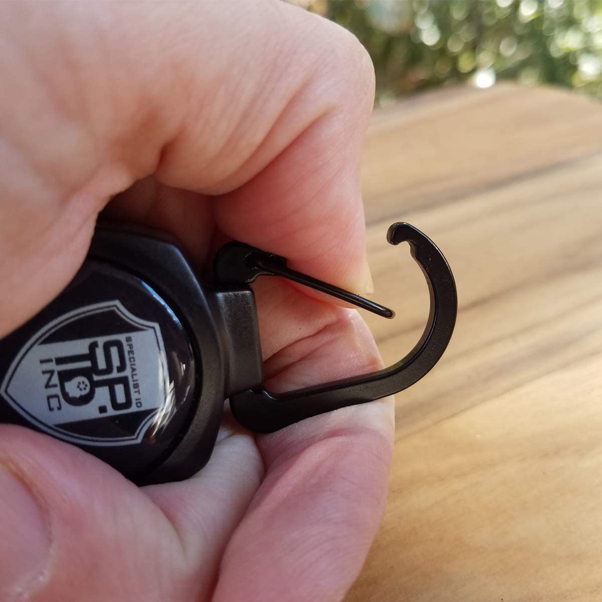 Close-up of a hand holding a SPID Key-Bak SIDEKICK Heavy Duty Retractable Carabiner Badge Reel with ID Holder Strap & Keychain with a metal hook, partially extended against a wooden surface.