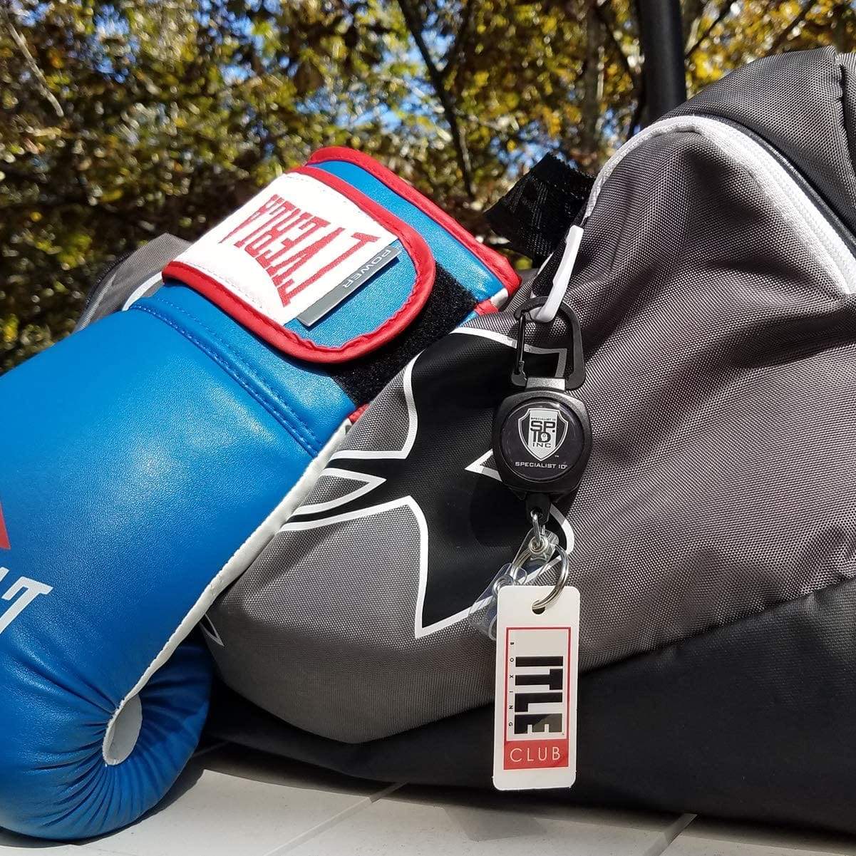 Close-up of a blue boxing glove resting on a gray Under Armour backpack with a Title Club gym lock attached, outdoors with tree leaves in the background. The bag also features an SPID Key-Bak SIDEKICK Heavy Duty Retractable Carabiner Badge Reel with ID Holder Strap & Keychain for securing accessories.