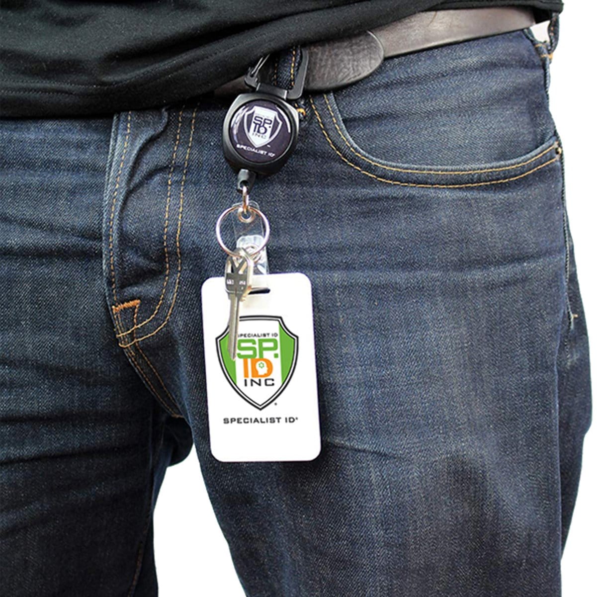 A person wearing jeans with a belt has an ID badge reel clipped to their waistband, displaying a "Specialist ID Inc." badge with a green and yellow shield logo. The SPID Key-Bak SIDEKICK Heavy Duty Retractable Carabiner Badge Reel with ID Holder Strap & Keychain, paired with a Kevlar retractable cord, ensures durability and security.
