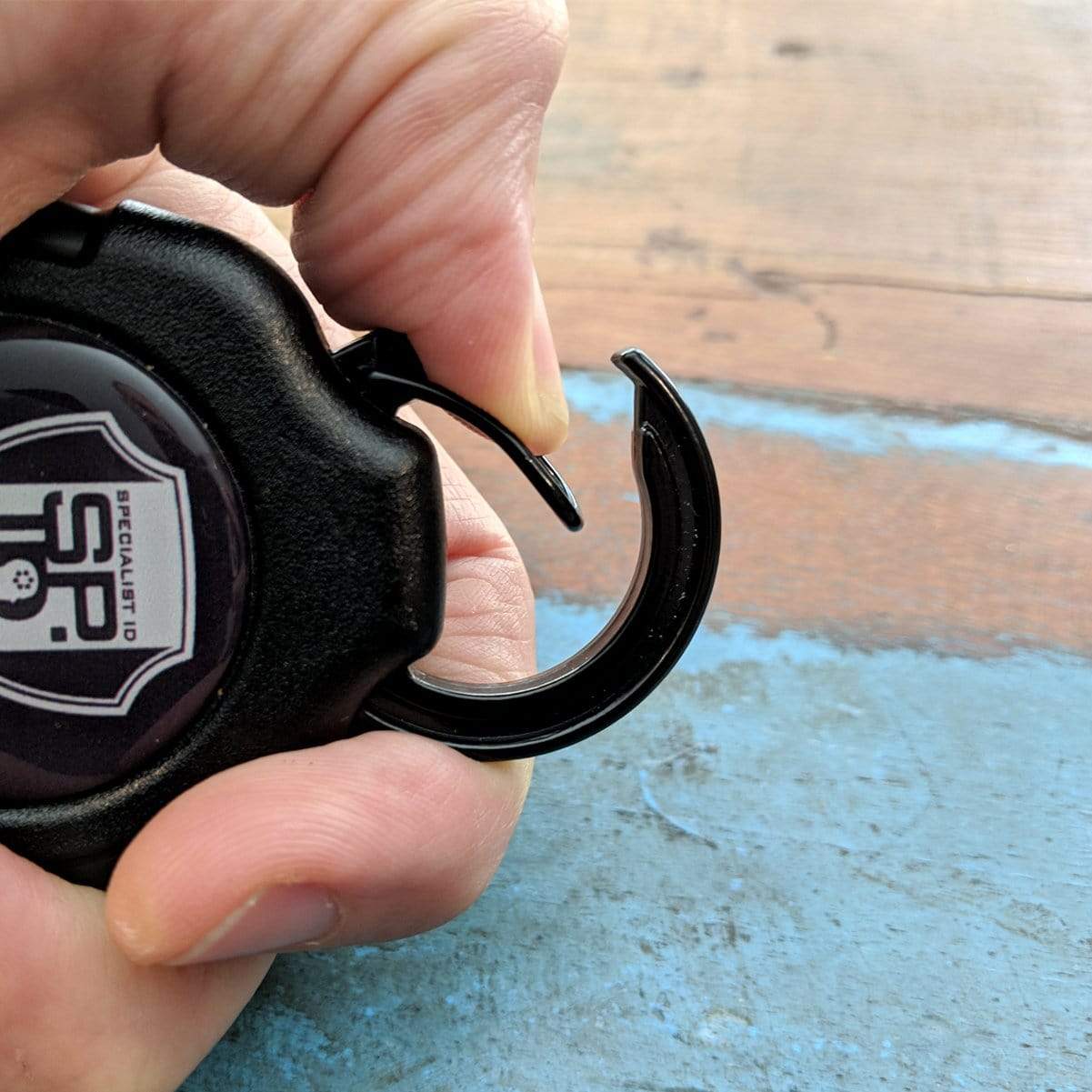A hand is holding a Heavy Duty Retractable Ratchit Keychain Tether Reel for Multiple Keys with Clip and Locking Feature with a black plastic hook, a logo on the clip, and a heavy-duty key reel.