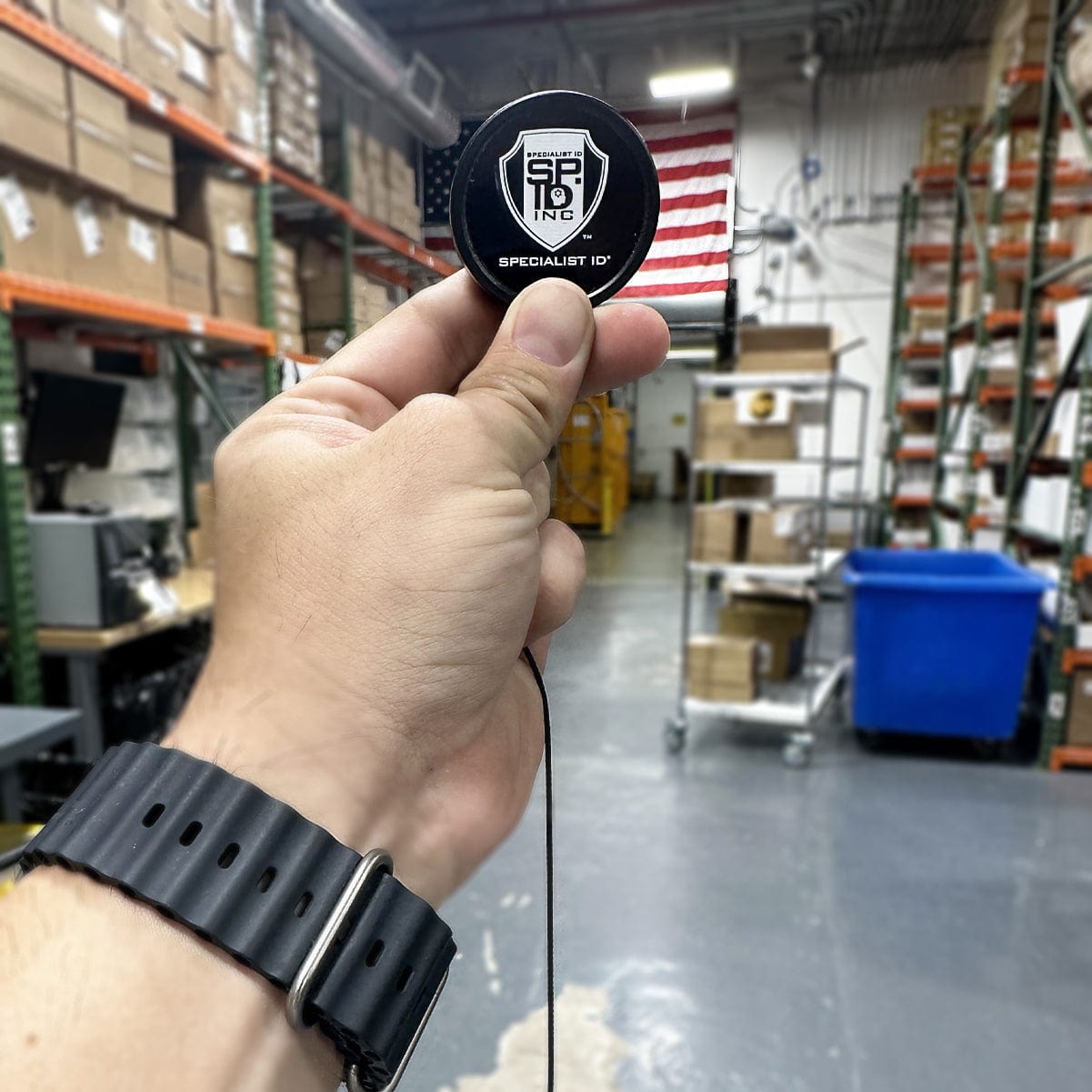 A hand holding a Heavy Duty Badge Reel w/ Metal Swivel Belt Clip & Key Ring with the logo "SP19 INC Specialist ID" in a warehouse setting with stacked boxes and shelving. The sturdy metal swivel belt clip ensures secure attachment to your work attire.
