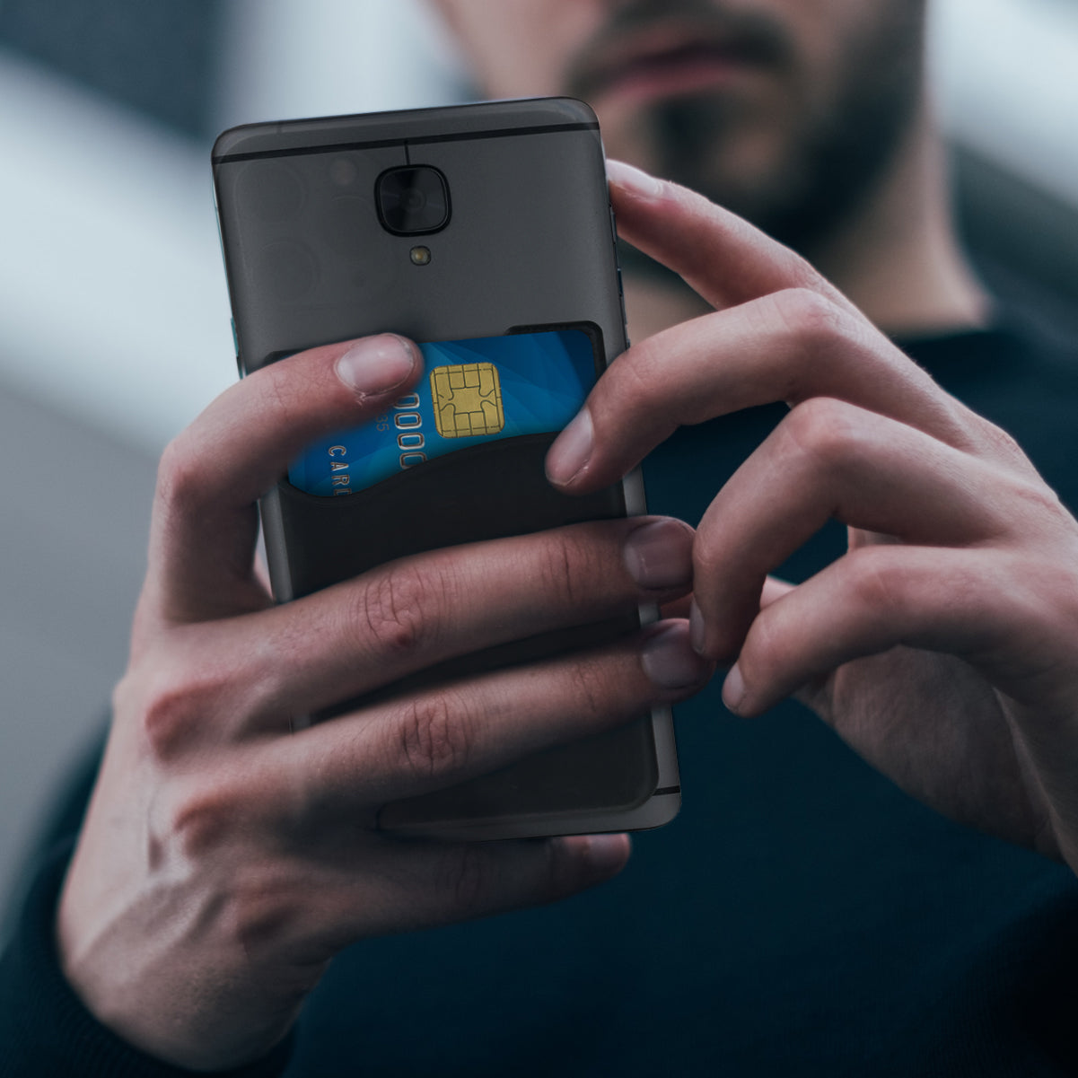 A person holds a phone with one hand and inserts a blue credit card into the Silicone Cell Phone Wallet (SPID-050X) attached to the case with adhesive backing.