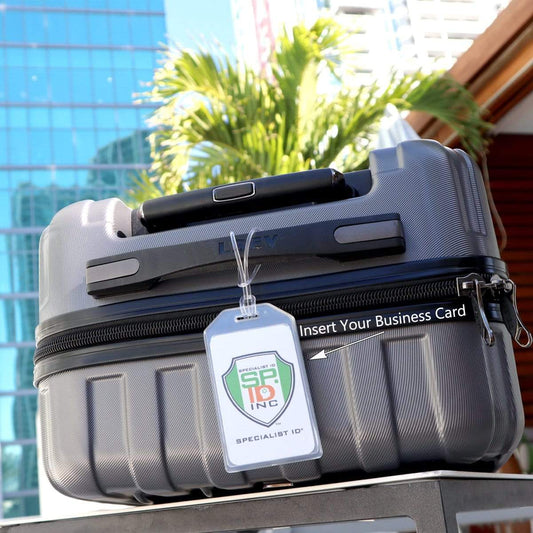 A silver hard-shell suitcase with Heavy Duty Luggage Tags with Plastic Loops Included - Window for Business Card or Included Insert (1840-620X) is positioned outdoors, framed by a modern glass building and a palm tree in the background.