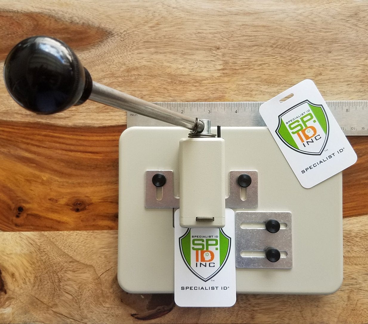 A Heavy Duty Table Top Slot Punch With Adjustable Guides (P/N SPID-9620) with a black handle, metal base, and two sample ID badges displaying the "Specialist ID" logo, placed on a wooden surface with a ruler next to it.