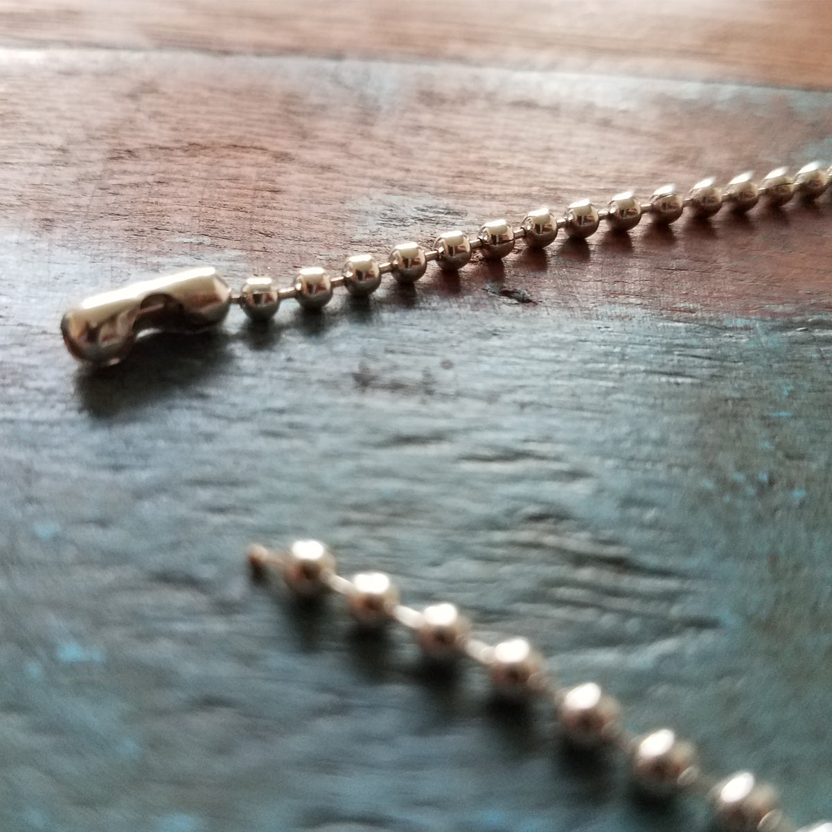 Close-up of a broken 2125-1000 Nickel-Plated Steel 24" Beaded Neck Chain with a clasp, lying on a wooden surface with one end of the chain separated from the other.