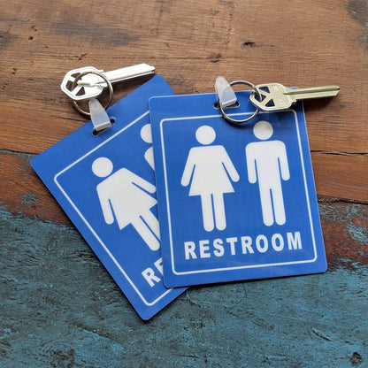 Two Unisex Restroom Pass Keychain - Bathroom Tag with Key Chain Ring - Heavy Duty Large Passes for Unisex & Family Restrooms with Key Holder (Sold in 2 Pack) (SPID-9840-Blue) rest on a wooden surface. The durable pass keychains feature unisex restroom symbols for male and female, labeled "RESTROOM.
