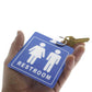 A hand holds a blue Unisex Restroom Pass Keychain - Bathroom Tag with Key Chain Ring - Heavy Duty Large Passes for Unisex & Family Restrooms with Key Holder (Sold in 2 Pack) (SPID-9840-Blue) featuring male and female symbols and the word "RESTROOM," attached to a key on a keyring, serving as a unisex restroom pass.