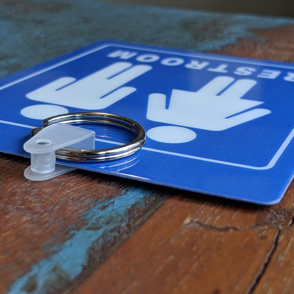 Close-up of a blue Unisex Restroom Pass Keychain - Bathroom Tag with Key Chain Ring - Heavy Duty Large Passes for Unisex & Family Restrooms with Key Holder (Sold in 2 Pack) (SPID-9840-Blue) with male and female symbols attached to a durable key holder, laying flat on a wooden surface.