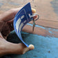 A hand holds a durable blue piece of plastic, featuring two human icons, attached to a keyring and a key. The background showcases a wooden surface with blue paint. This versatile item serves as a Unisex Restroom Pass Keychain - Bathroom Tag with Key Chain Ring - Heavy Duty Large Passes for Unisex & Family Restrooms with Key Holder (Sold in 2 Pack) (SPID-9840-Blue), perfect for any setting.