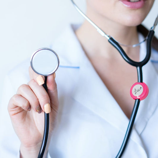 A person in a white coat holds up a stethoscope with an Antimicrobial Stethoscope ID Tag - Hand Write Your Name on Label to Personalize - Adjustable Tube Size to Fit Most Steths, emphasizing safety and personalization.