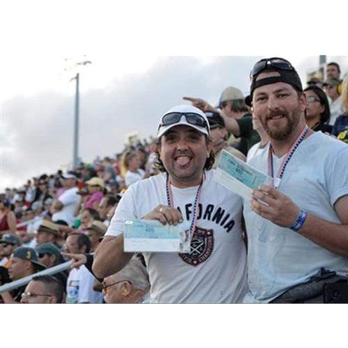 Two men standing and smiling in a crowded stadium, holding tickets. One man is wearing a white t-shirt with "California" on it, while the other has a black cap, blue long sleeve shirt, and an oversized Vertical Oversized 4X7 Vinyl ID Badge & Ticket Holder (XL47.5V) around his neck.