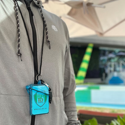 A person wearing a grey jacket with a neck lanyard holds a blue Witz See It Safe Waterproof ID Badge Holder (P/N 004) that says "Specialist ID." The crush resistant plastic case provides durability. In the background, you can see a pool with green and yellow striped tubes.