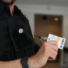 A person in a black uniform showcases an ID card connected to the Custom Compact Key-Bak Retractable Carabiner ID Badge Reel with Key Ring and Your Logo, enhancing durability. Sold by Specialist ID.
