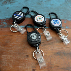 Four **Custom Compact Key-Bak Retractable Carabiner ID Badge Reels with Key Rings and Your Logo** are displayed on a wooden surface. Each Key-Bak holder features a different design or logo on its front, complete with durable retractable Kevlar cords.