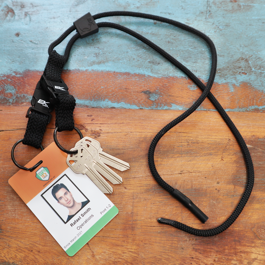 An EK Black Lanyard With Two Detachable Key Rings (10046) by EK USA, adorned with keys and a badge labeled "Rafael Smith, Operations, Plant 12," lies on a wooden surface. Sold by Specialist ID.