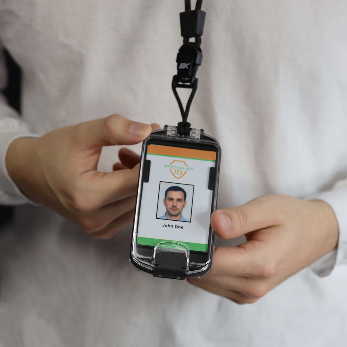 A person in a white shirt holds an EK USA Guardian RFID Blocking Hard Shell Badge Holder with Detachable Lanyard (10935), displaying an ID badge featuring a photo and the name “John Doe.” The badge is securely encased, providing both protection and professionalism.