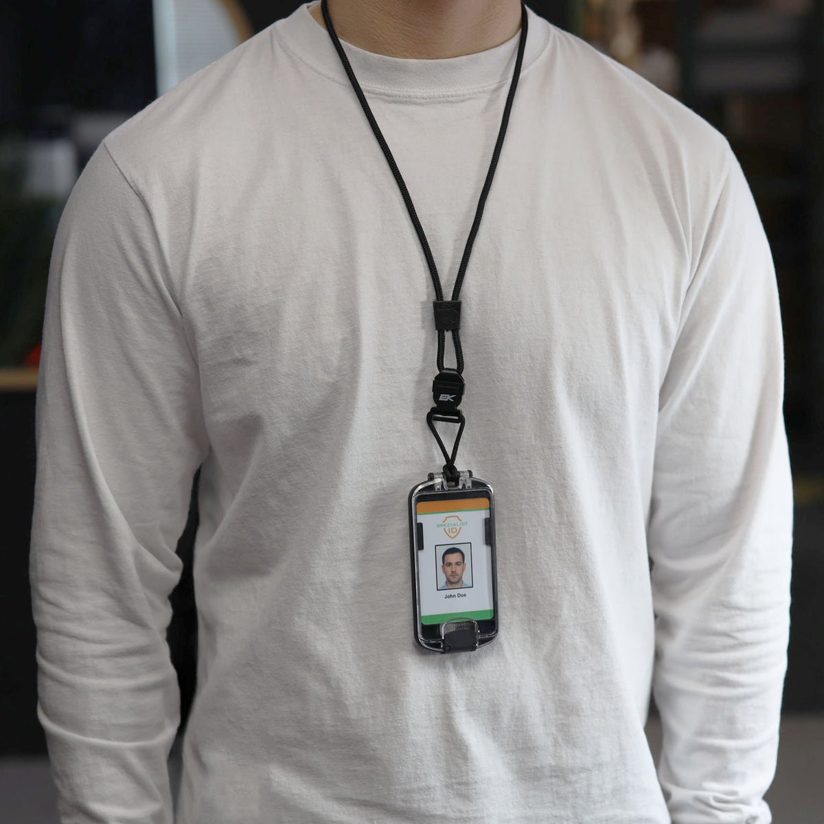 An individual wearing a white long-sleeve shirt stands indoors with an ID badge—featuring a photo and text—securely displayed in the EK USA Guardian RFID Blocking Hard Shell Badge Holder with Detachable Lanyard (10935).