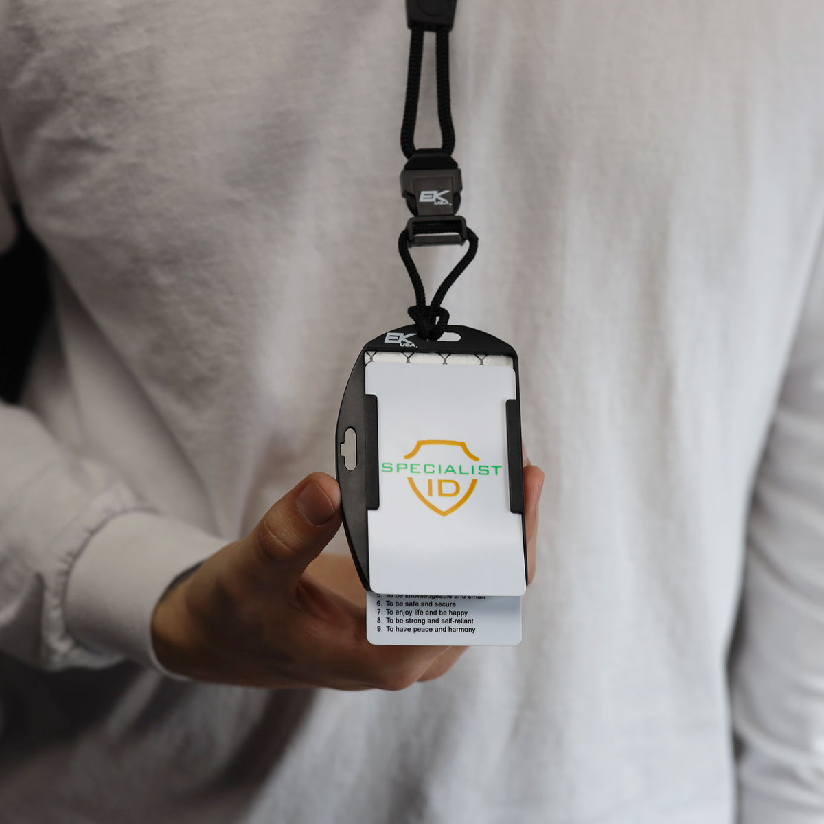 A person wearing a white shirt holds the EK USA Patriot RFID Blocking Dual Sided Badge Holder with Detachable Lanyard (10943), which displays an ID card labeled “SPECIALIST ID” with a shield logo.