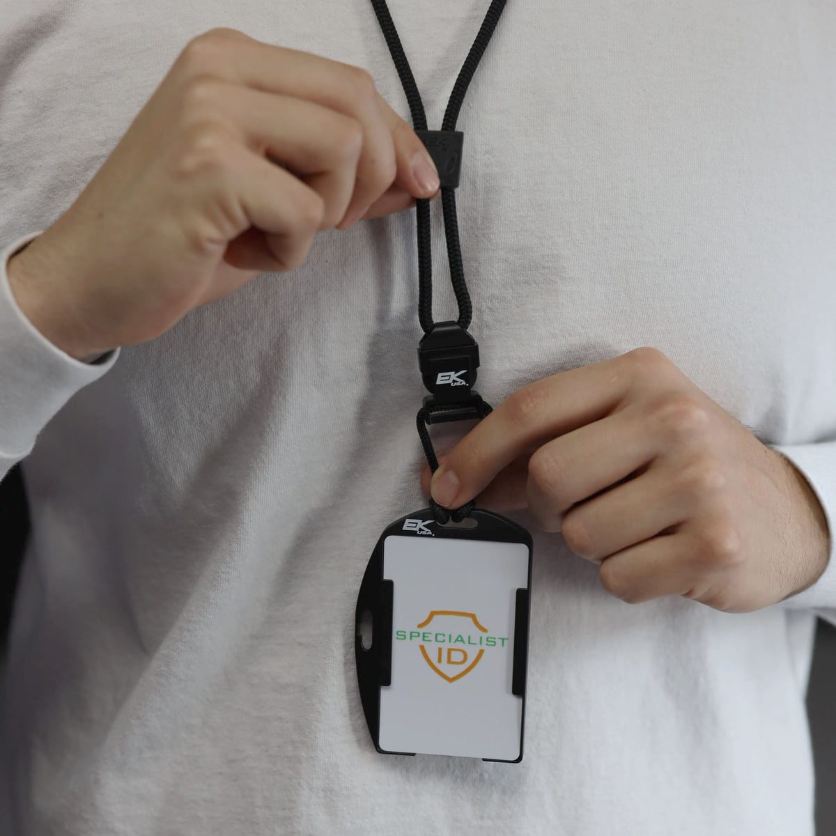 A person wearing a white long-sleeve shirt holds the EK USA Patriot RFID Blocking Dual Sided Badge Holder with Detachable Lanyard (10943), which displays the words “Specialist ID” and a shield logo.