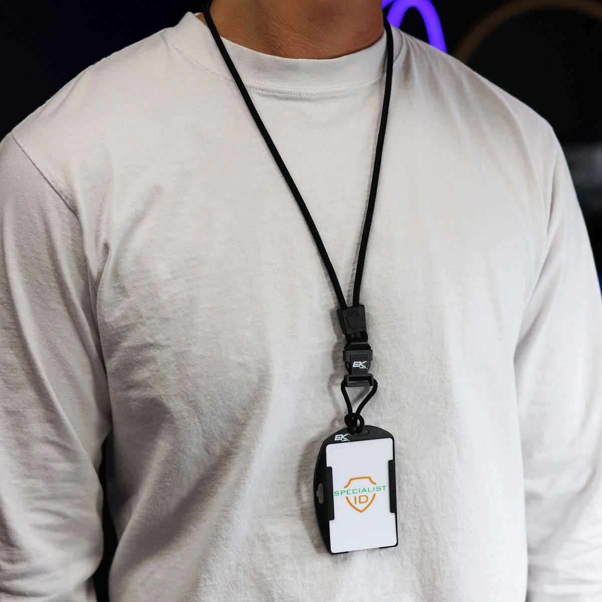 A person wearing a plain white long-sleeve shirt with an EK USA Patriot RFID Blocking Dual Sided Badge Holder with Detachable Lanyard (10943) and an ID badge around their neck, standing against a dark background.