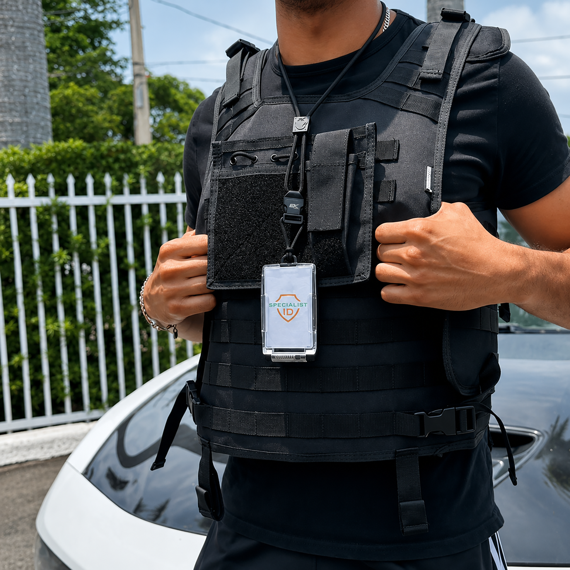 A person wearing a black tactical vest holds the EK USA One Hander RFID Blocking Badge Holder with Detachable Lanyard (10983) in front of a white car and a metal fence.