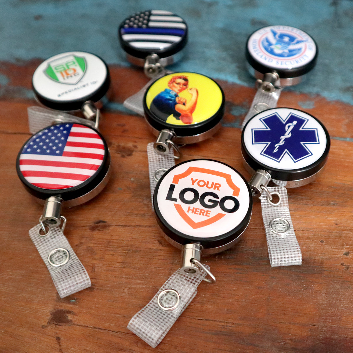 Heavy Duty Custom Printed Badge Reel With Steel Cable 2120-3305 - Add Your Logo with various designs including flags, medical symbols, and a customized logo, arranged on a wooden surface to boost brand awareness.